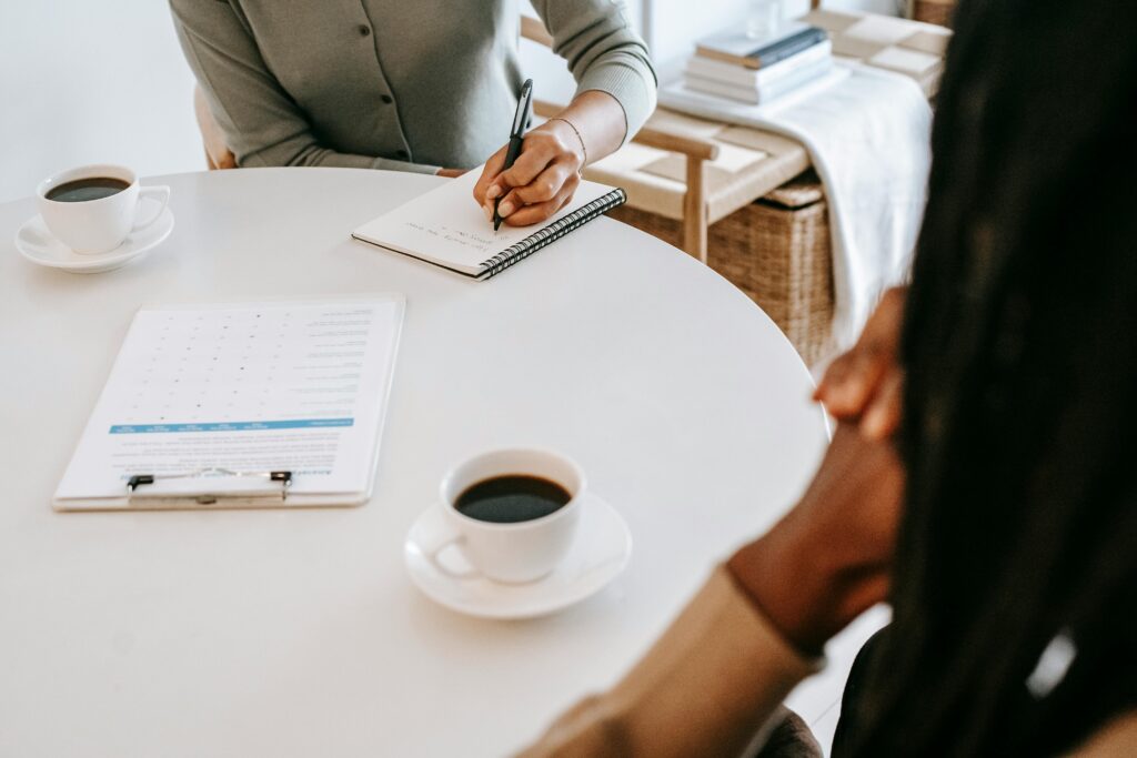 Get in Touch From above crop multiracial female interviewer or psychologist in formal wear asking questions and taking notes in planner while talking to black man and sitting together at table with coffee